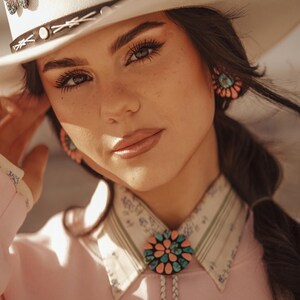 May include: A woman wearing a white cowboy hat with a butterfly pin, a pink shirt with a turquoise and pink bolo tie, and turquoise earrings.