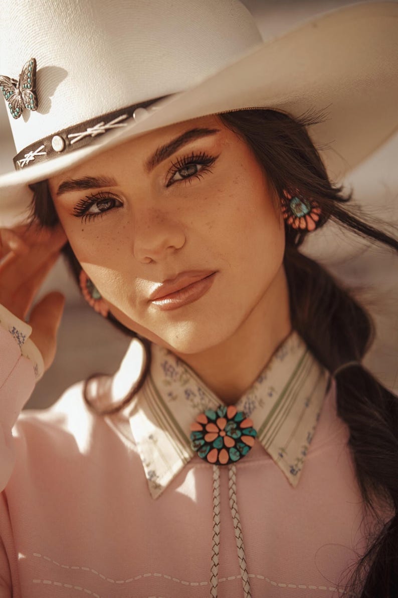 May include: A woman wearing a white cowboy hat with a butterfly pin, a pink shirt with a turquoise and pink bolo tie, and turquoise earrings.