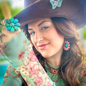 May include: A woman wearing a brown hat with a turquoise butterfly pin, a turquoise flower ring, and pink flower earrings. She is wearing a green floral blouse.