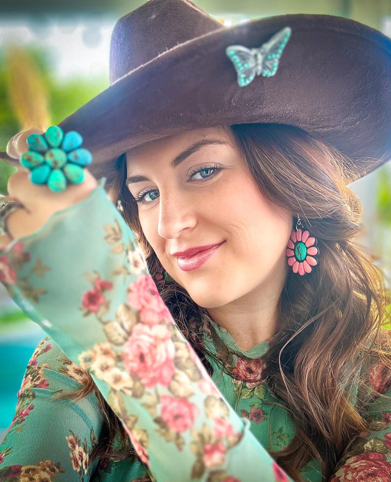 May include: A woman wearing a brown hat with a turquoise butterfly pin, a turquoise flower ring, and pink flower earrings. She is wearing a green floral blouse.