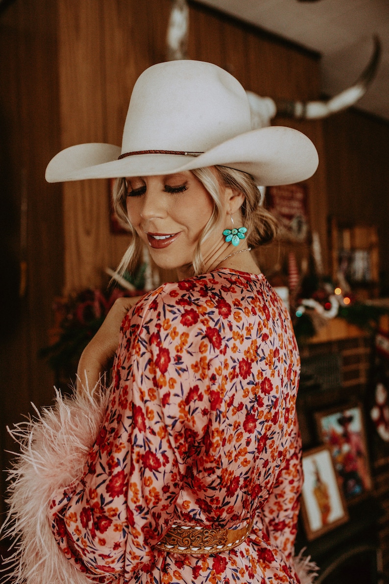Pode incluir: Uma mulher usando um chap&eacute;u de cowboy branco, um vestido com estampa floral e brincos turquesa. O vestido tem flores vermelhas em um fundo rosa. As mangas s&atilde;o adornadas com penas rosa. Um cinto marrom com fivela decorativa afivela a cintura.