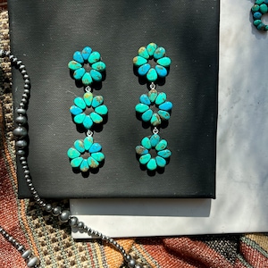 May include: Turquoise flower earrings with three tiers of turquoise petals. The earrings are displayed on a black surface, alongside a matching turquoise pendant necklace and a beaded necklace. The turquoise has brown matrix.