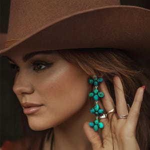 May include: A brown cowboy hat and turquoise earrings. The earrings are composed of clusters of turquoise stones. The hat has a brown band. The image is a close-up of a person wearing the hat and earrings.