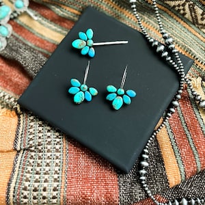 Pode incluir: Um conjunto de joias de turquesa, incluindo uma presilha de cabelo e brincos, exibido em uma superf&iacute;cie preta. Um colar com contas de prata e um colar de turquesa tamb&eacute;m s&atilde;o vis&iacute;veis. As joias s&atilde;o colocadas em um fundo t&ecirc;xtil tecido colorido.