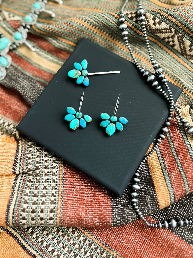 Pode incluir: Um conjunto de joias de turquesa, incluindo uma presilha de cabelo e brincos, exibido em uma superf&iacute;cie preta. Um colar com contas de prata e um colar de turquesa tamb&eacute;m s&atilde;o vis&iacute;veis. As joias s&atilde;o colocadas em um fundo t&ecirc;xtil tecido colorido.