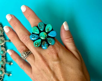 May include: A large turquoise ring with a flower design. The ring features multiple teardrop-shaped turquoise stones in shades of blue and green, set on a black base. A silver ring with a diamond-like stone is also visible.