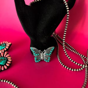 May include: A silver chain necklace with turquoise butterfly pendant on a pink background.