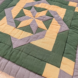 Può includere: Un tappetino quadrato trapuntato con un design geometrico a stella. Il tappetino presenta un motivo a stella centrale in giallo e grigio, circondato da blocchi verdi e grigi. I bordi sono rifiniti con un bordo grigio, creando un aspetto accogliente e fatto a mano.