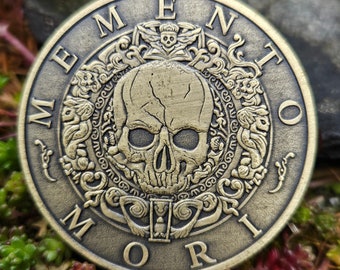 Op de afbeelding: Een ronde, messingkleurige munt met een schedel en kruisbeenderen ontwerp. De munt heeft de woorden "Memento Mori" gegraveerd rond de rand.