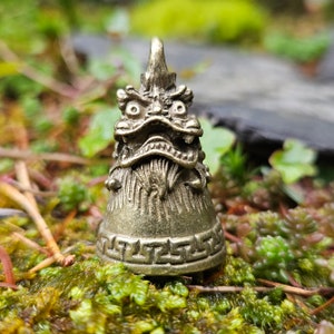 Puede incluir: Una pequeña campana de metal dorada con un diseño de dragón detallado. La campana está sentada sobre un lecho de musgo verde.