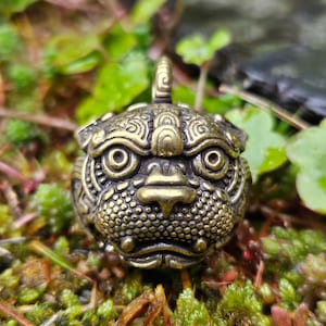 Puede incluir: Colgante de latón de estilo antiguo con forma de cabeza de animal estilizada. El colgante presenta detalles intrincados, incluyendo superficies texturizadas y un lazo para colgar. El diseño sugiere una estética vintage o artesanal.