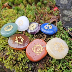 Op de afbeelding: Zeven gepolijste edelstenen in verschillende kleuren, waaronder groen, wit, paars, blauw, bruin en oranje. Elke steen heeft een goudkleurig chakra-symbool. De stenen zijn gerangschikt op een bed van groen mos.
