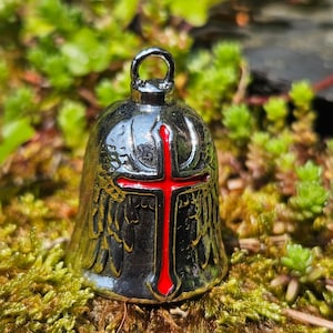 May include: A silver bell with a red cross design. The bell has wing patterns and a loop at the top. The background is a blurred view of green plants and moss.