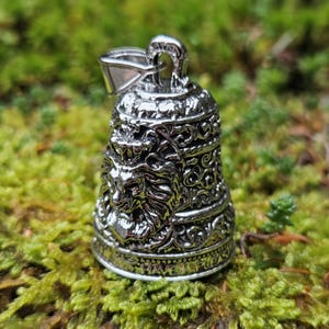 Puede incluir: Colgante de campana plateada con un diseño detallado de cabeza de león y adornos de volutas. La campana está suspendida de un pequeño lazo y descansa sobre un lecho de musgo verde.
