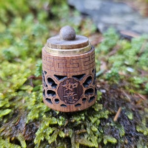 Może przedstawiać: Mały drewniany pojemnik z złotą obwódką i rzeźbionym wzorem. Pojemnik ma okrągłą pokrywkę z gałką na górze. Pojemnik stoi na łóżku z zielonego mchu.