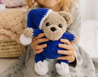 May include: A handmade, plush teddy bear in a blue and tan color scheme. The bear wears a blue nightcap with a white trim and white feet. The bear is held by a person.