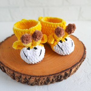 K&ouml;nnte beinhalten: Ein Paar gelbe gestrickte Babyschuhe mit braunen Bommeln und wei&szlig;en Garn-Gesichtern. Die Schuhe sind in Form von Giraffen und liegen auf einer Holzoberfl&auml;che.