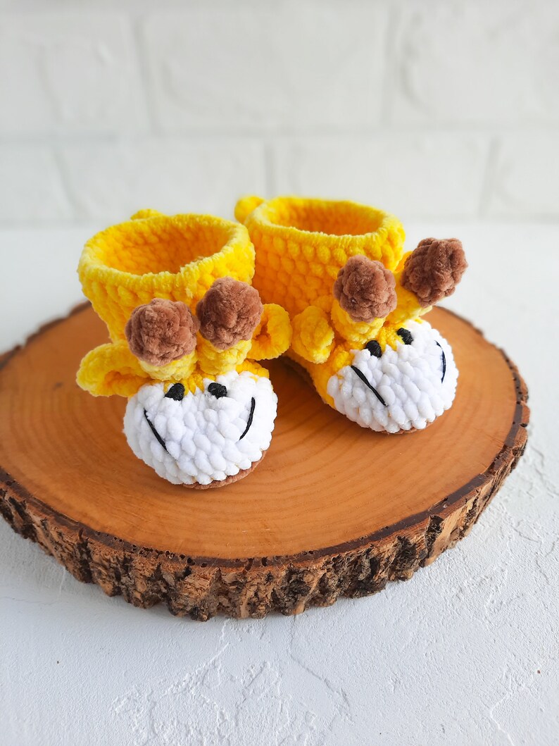 K&ouml;nnte beinhalten: Ein Paar gelbe gestrickte Babyschuhe mit braunen Bommeln und wei&szlig;en Garn-Gesichtern. Die Schuhe sind in Form von Giraffen und liegen auf einer Holzoberfl&auml;che.