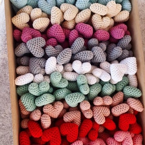 Peut inclure: Une collection de cœurs au crochet de différentes couleurs, notamment rouge, rose, vert, gris, blanc et bleu. Les cœurs sont disposés en rangées, les plus petits en bas et les plus grands en haut.