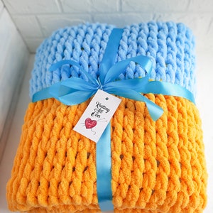 K&ouml;nnte beinhalten: Eine gestrickte Wolldecke in Orange und Hellblau mit einem blauen Band darum gebunden. Die Decke hat ein Etikett mit der Aufschrift "Knitting All Over".