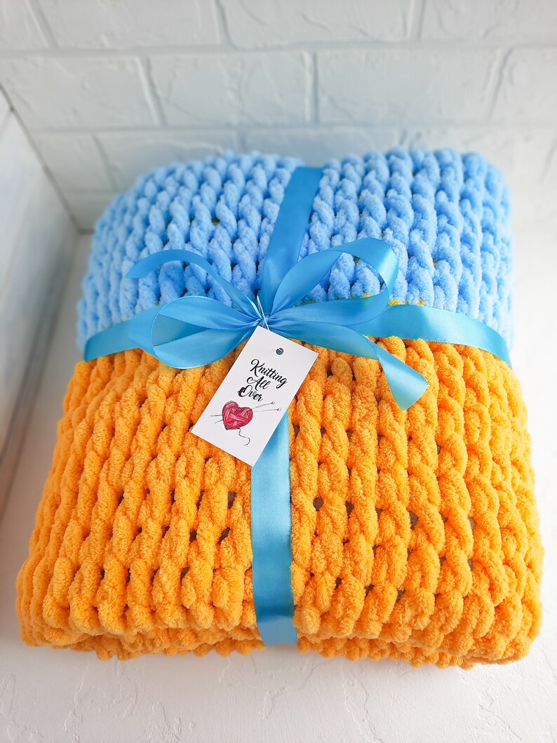K&ouml;nnte beinhalten: Eine gestrickte Wolldecke in Orange und Hellblau mit einem blauen Band darum gebunden. Die Decke hat ein Etikett mit der Aufschrift "Knitting All Over".