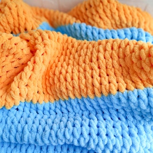 K&ouml;nnte beinhalten: Eine weiche, dicke Strickdecke in blau und orange. Die Decke ist aus dickem Garn gefertigt und hat eine flauschige Textur.