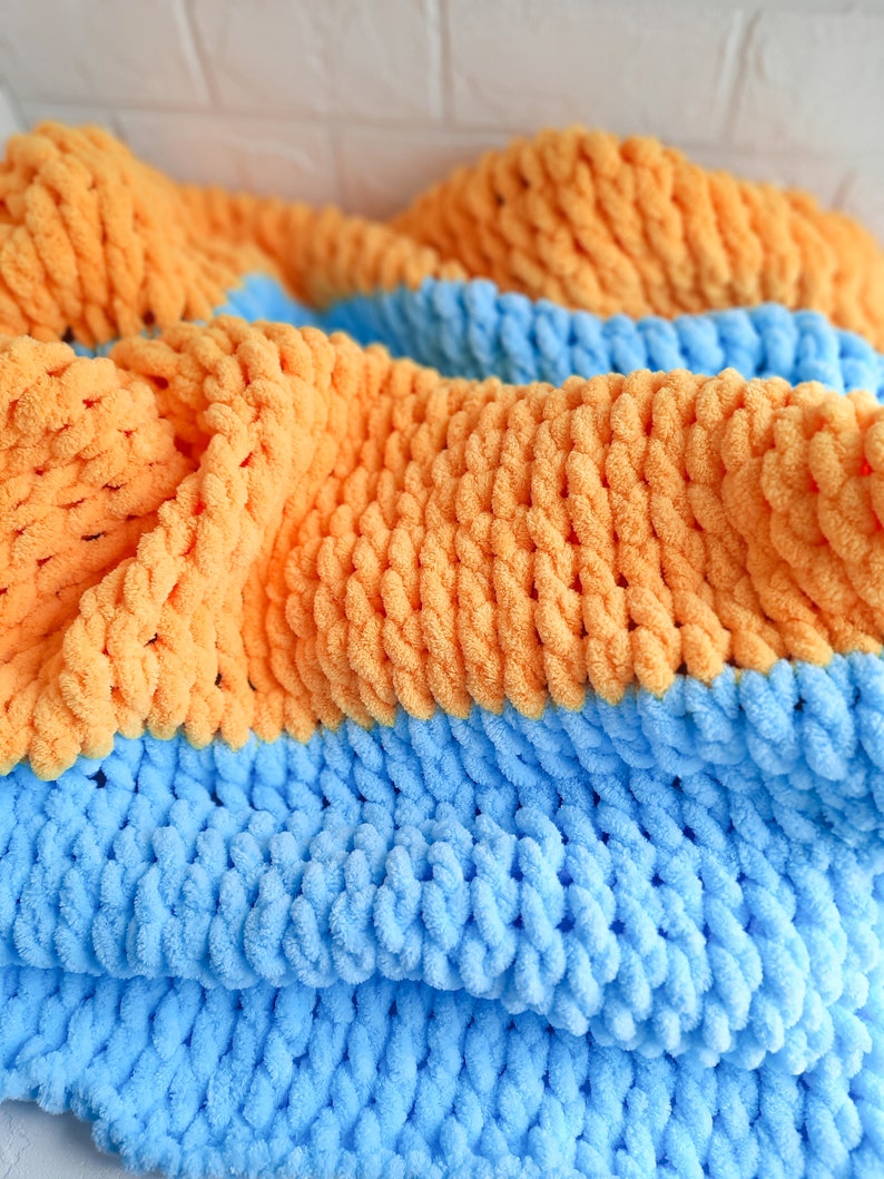 K&ouml;nnte beinhalten: Eine weiche, dicke Strickdecke in blau und orange. Die Decke ist aus dickem Garn gefertigt und hat eine flauschige Textur.