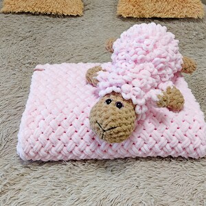 Peut inclure: Une couverture en crochet rose avec un jouet en crochet agneau brun et rose sur le dessus. La couverture et le jouet sont sur une surface moelleuse brun clair.