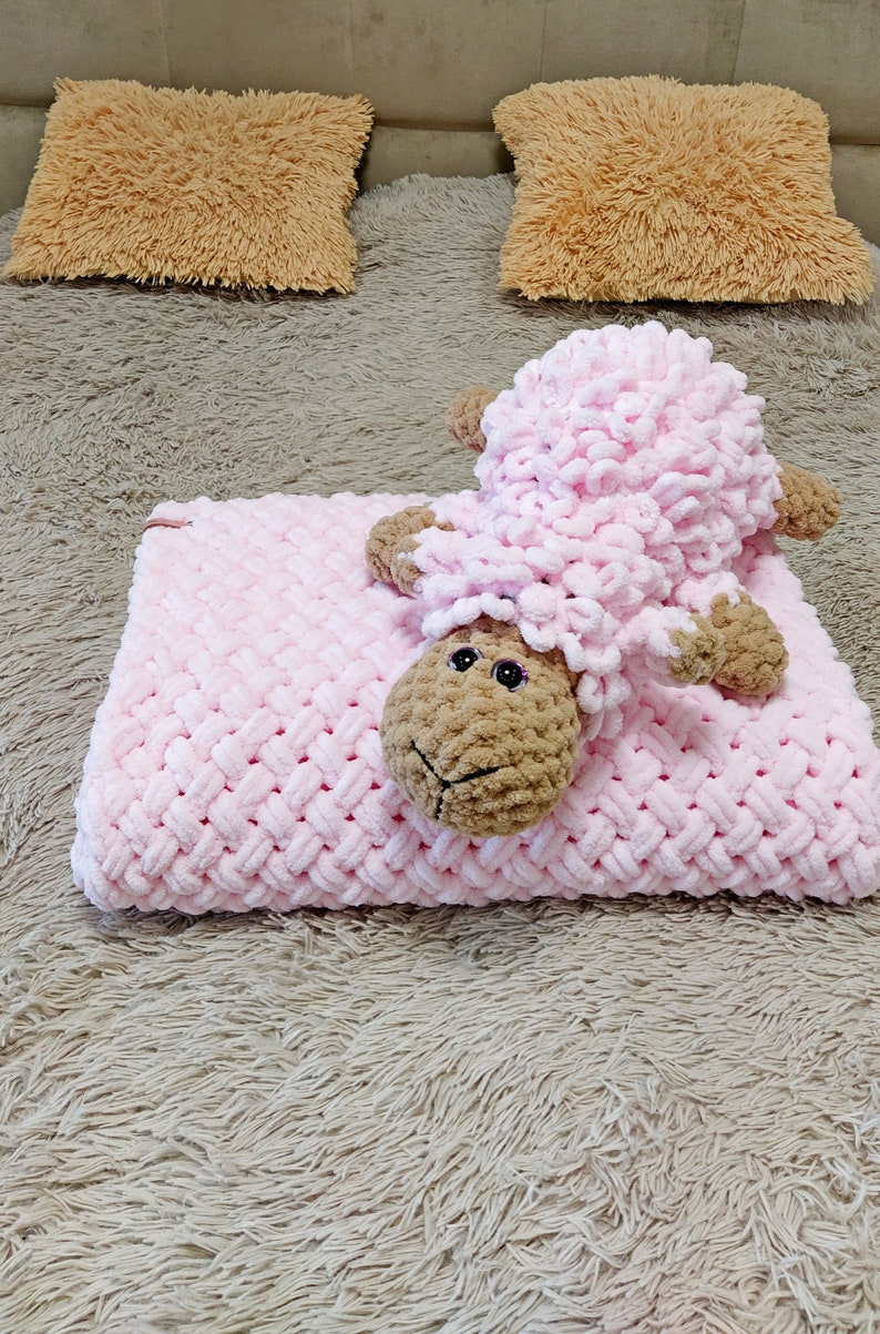 Peut inclure: Une couverture en crochet rose avec un jouet en crochet agneau brun et rose sur le dessus. La couverture et le jouet sont sur une surface moelleuse brun clair.