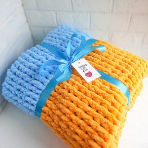 K&ouml;nnte beinhalten: Eine blaue und orange gestrickte Decke, die mit einem blauen Band und einem Geschenkanh&auml;nger mit der Aufschrift "Knitting All Over" gebunden ist.