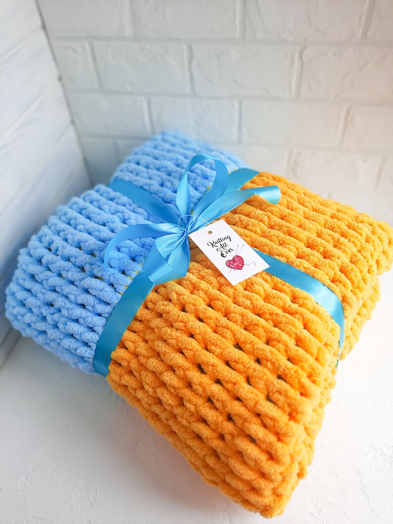K&ouml;nnte beinhalten: Eine blaue und orange gestrickte Decke, die mit einem blauen Band und einem Geschenkanh&auml;nger mit der Aufschrift "Knitting All Over" gebunden ist.