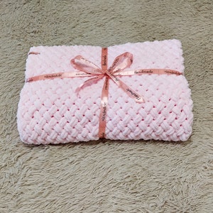 Peut inclure: Une couverture pour b&eacute;b&eacute; en crochet rose avec un ruban rose nou&eacute; autour. Le ruban porte l'inscription "Handmade" r&eacute;p&eacute;t&eacute;e plusieurs fois.