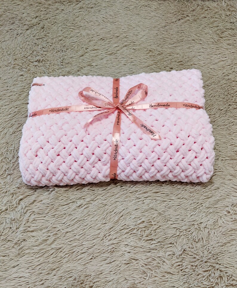 Peut inclure: Une couverture pour b&eacute;b&eacute; en crochet rose avec un ruban rose nou&eacute; autour. Le ruban porte l'inscription "Handmade" r&eacute;p&eacute;t&eacute;e plusieurs fois.