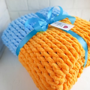 K&ouml;nnte beinhalten: Eine weiche, gestrickte Decke in einer Kombination aus leuchtendem Orange und Hellblau. Die Decke ist mit einem blauen Band gebunden und hat ein kleines Etikett mit dem Text "Handmade with Love".