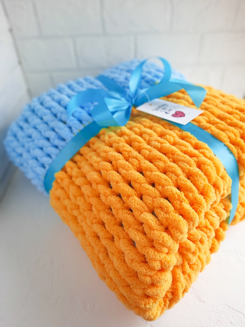 K&ouml;nnte beinhalten: Eine weiche, gestrickte Decke in einer Kombination aus leuchtendem Orange und Hellblau. Die Decke ist mit einem blauen Band gebunden und hat ein kleines Etikett mit dem Text "Handmade with Love".