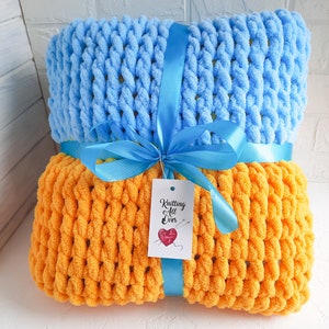 K&ouml;nnte beinhalten: Zwei gestrickte Decken, eine hellblau und eine orange, liegen &uuml;bereinander. Sie sind mit einem blauen Band und einem Etikett mit der Aufschrift "Knitting All Over" zusammengebunden.