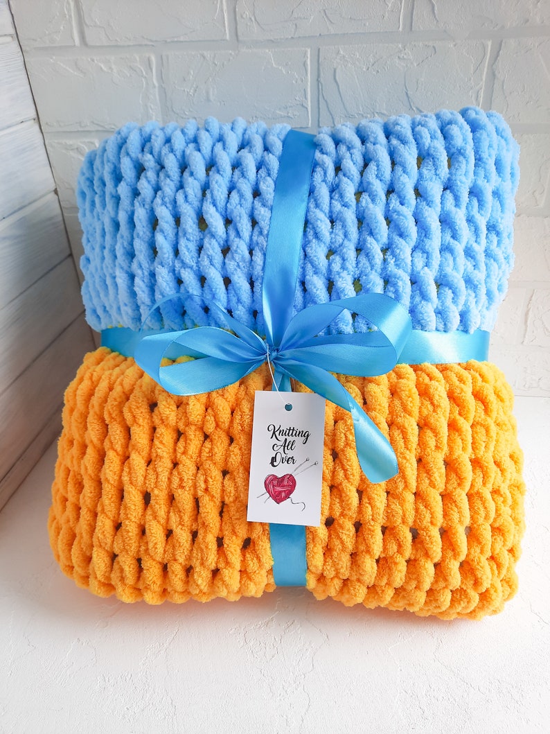 K&ouml;nnte beinhalten: Zwei gestrickte Decken, eine hellblau und eine orange, liegen &uuml;bereinander. Sie sind mit einem blauen Band und einem Etikett mit der Aufschrift "Knitting All Over" zusammengebunden.