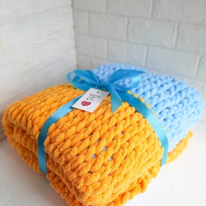 K&ouml;nnte beinhalten: Zwei gestrickte Decken, eine orange und eine hellblau, sind zusammengelegt und mit einem blauen Band gebunden. Ein kleines Etikett mit dem Text "Knitting with Love" ist am Band befestigt.