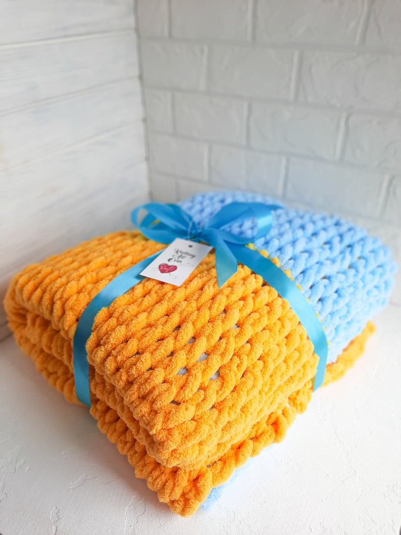 K&ouml;nnte beinhalten: Zwei gestrickte Decken, eine orange und eine hellblau, sind zusammengelegt und mit einem blauen Band gebunden. Ein kleines Etikett mit dem Text "Knitting with Love" ist am Band befestigt.