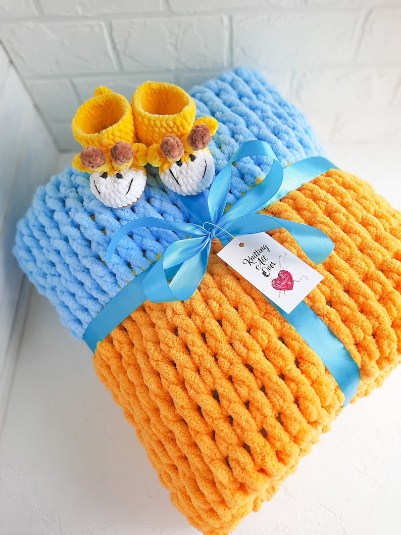 K&ouml;nnte beinhalten: Eine gestrickte Babydecke in Blau und Orange mit einem blauen Band und einem Paar gelber gestrickter Babyschuhe mit Giraffenk&ouml;pfen. Die Decke ist mit einem blauen Band und einem Etikett mit der Aufschrift "Knitting Over" gebunden.