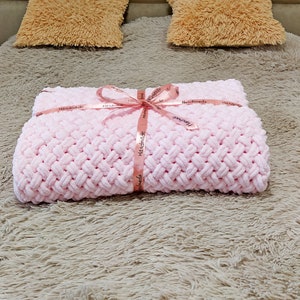 Peut inclure: Une couverture en tricot rose avec une texture tress&eacute;e, attach&eacute;e avec un ruban rose et le mot "Handmade" imprim&eacute; sur le ruban. La couverture est sur une surface moelleuse, brun clair.