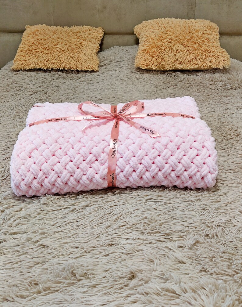 Peut inclure: Une couverture en tricot rose avec une texture tress&eacute;e, attach&eacute;e avec un ruban rose et le mot "Handmade" imprim&eacute; sur le ruban. La couverture est sur une surface moelleuse, brun clair.
