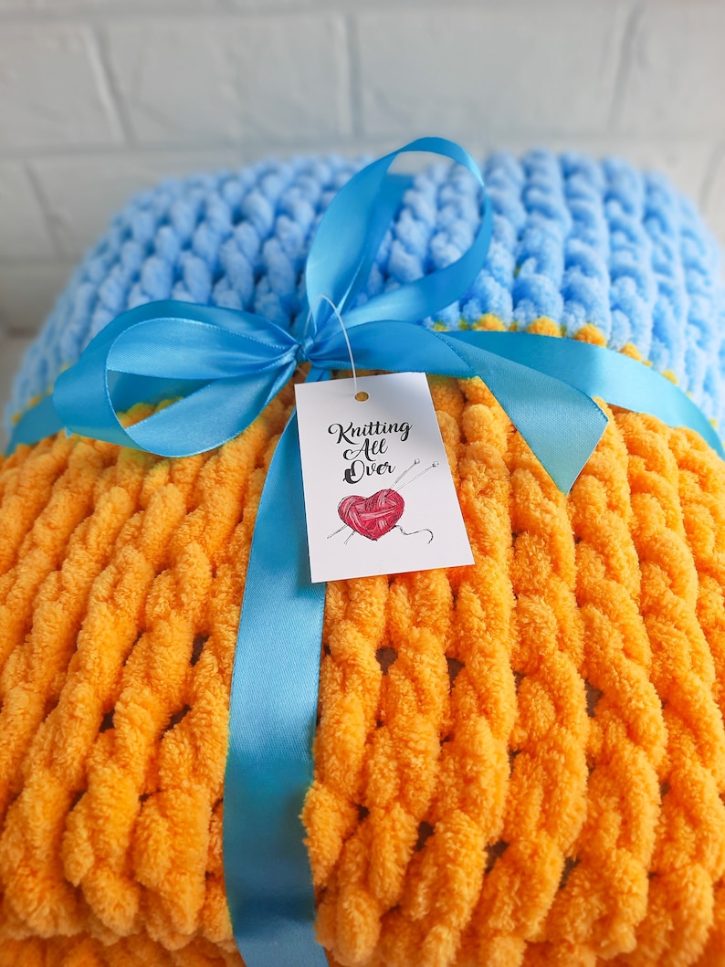 K&ouml;nnte beinhalten: Eine gelbe und blaue Strickdecke mit einem blauen Band darum gebunden. Die Decke hat ein Etikett mit der Aufschrift "Knitting All Over" mit einem Herzen und Stricknadeln.