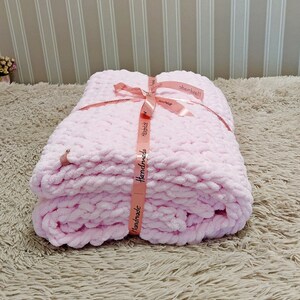 Peut inclure: Une couverture en crochet rose avec un ruban rose nou&eacute; autour. Le ruban porte l'inscription "Handmade".