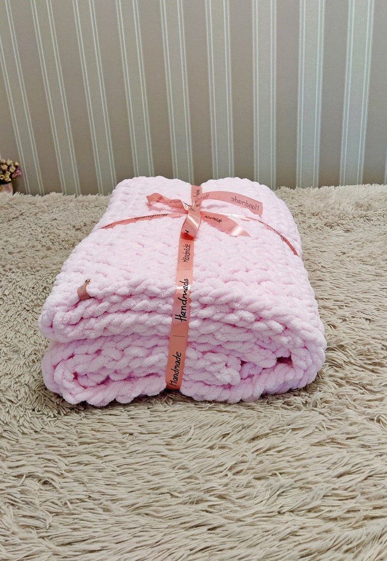 Peut inclure: Une couverture en crochet rose avec un ruban rose nou&eacute; autour. Le ruban porte l'inscription "Handmade".