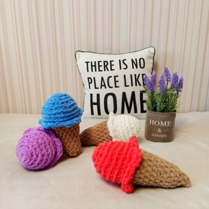 Peut inclure: Quatre c&ocirc;nes de cr&egrave;me glac&eacute;e en crochet de diff&eacute;rentes couleurs&nbsp;: violet, bleu, blanc et rouge. Les c&ocirc;nes sont pos&eacute;s sur une surface blanche et moelleuse. Un coussin avec le texte &laquo;&nbsp;Il n&rsquo;y a pas de place comme &agrave; la maison&nbsp;&raquo; se trouve en arri&egrave;re-plan.