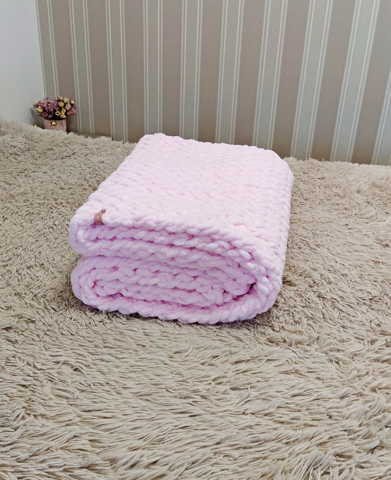 Peut inclure: Une couverture en tricot rose pli&eacute;e sur une surface moelleuse brun clair.