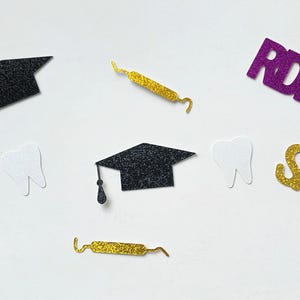 May include: A collection of glittery dental graduation-themed decorations. Includes black graduation caps, dental tools, white teeth, and the name "SARA" in gold glitter. Also features the letters "RDH" in purple glitter.