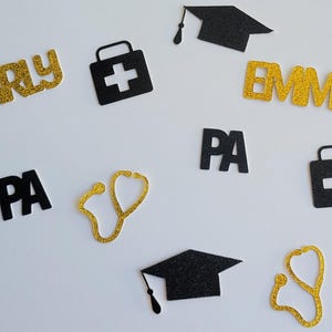 May include: Gold glitter letters spelling "KARLY" and "EMMA" and black glitter letters spelling "PA".  Black glitter graduation cap, stethoscope, and first aid kit shapes.