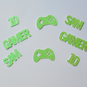 May include: Green glitter paper cutouts of the words "Gamer", "Sam", and the number "10". There are also two green glitter paper cutouts of a video game controller.